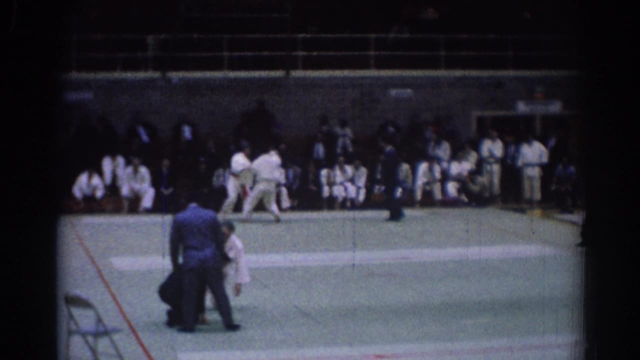 1967 Los Angeles frame showing a judo match in progress, with the referee standing near the athletes and spectators seated behind them.