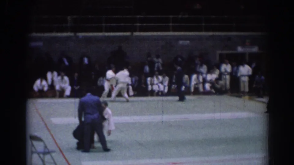 1967 Los Angeles frame showing a judo match in progress, with the referee standing near the athletes and spectators seated behind them.