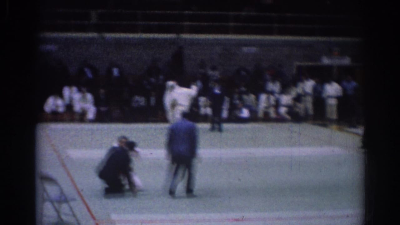 Soft 8mm frame from the 1967 Los Angeles judo competition, with motion-blurred athletes, a kneeling referee, and onlookers nearby.