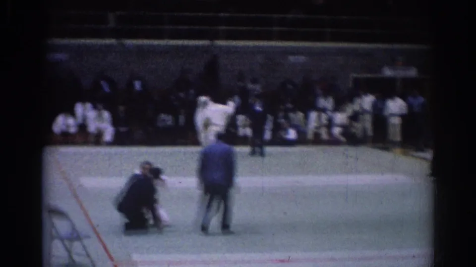Soft 8mm frame from the 1967 Los Angeles judo competition, with motion-blurred athletes, a kneeling referee, and onlookers nearby.