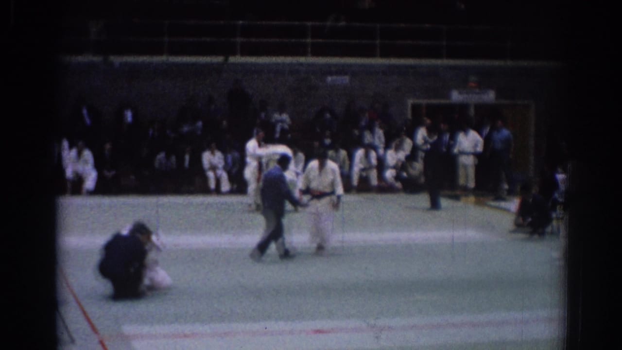 1967 Los Angeles home-movie frame with a referee crossing the mat, judo athletes nearby, and spectators seated along the gym wall.