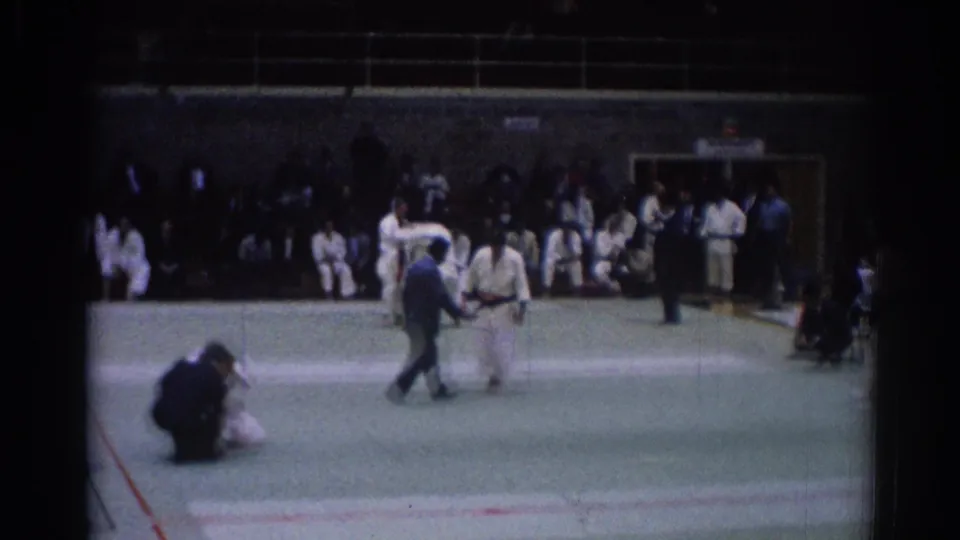 1967 Los Angeles home-movie frame with a referee crossing the mat, judo athletes nearby, and spectators seated along the gym wall.