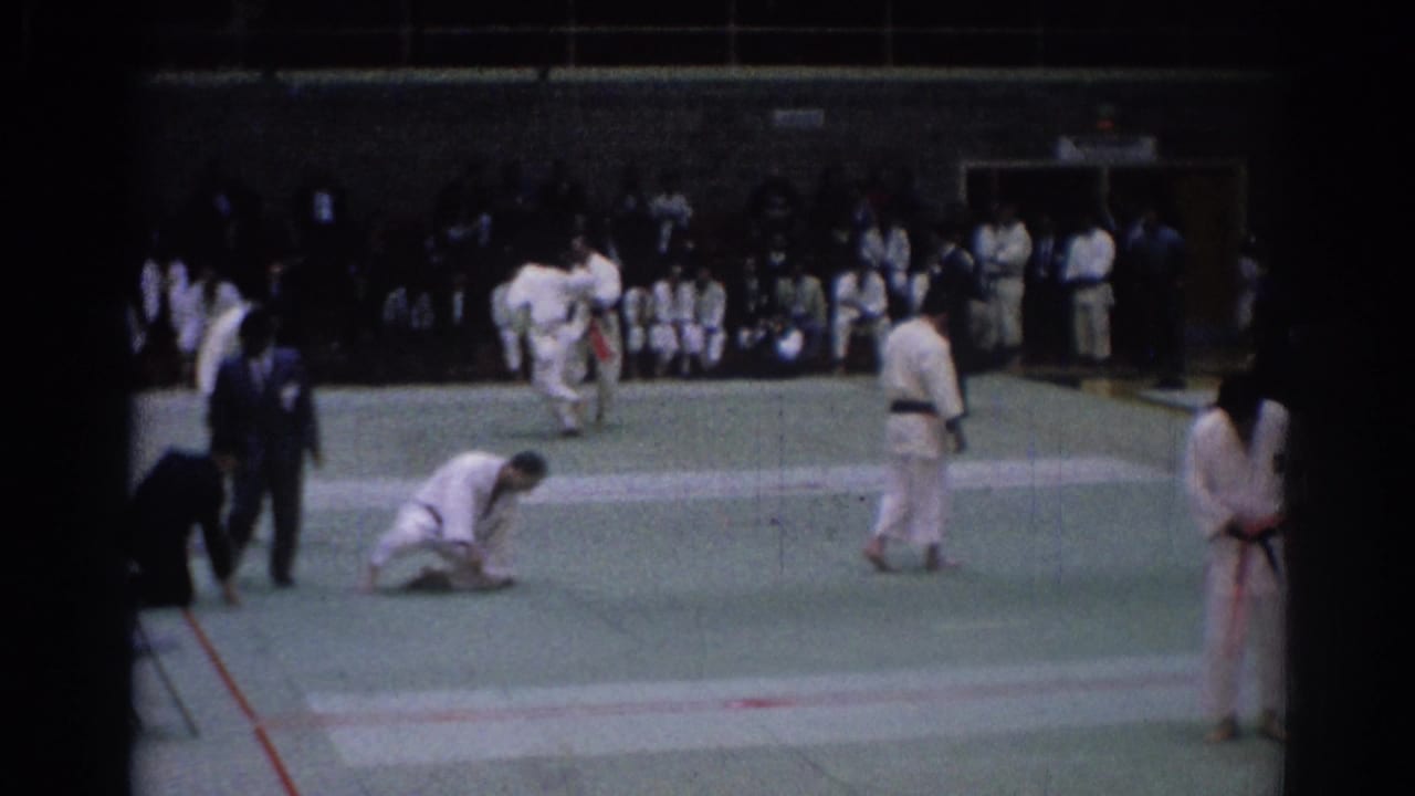 1967 Los Angeles karate competition frame with a kneeling athlete, standing competitors, judges, and sideline spectators.