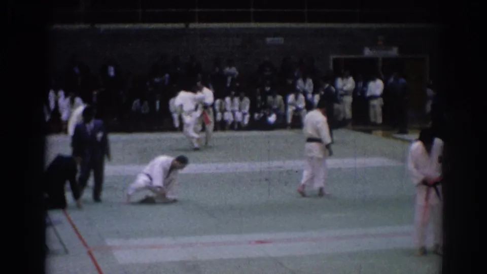 1967 Los Angeles karate competition frame with a kneeling athlete, standing competitors, judges, and sideline spectators.