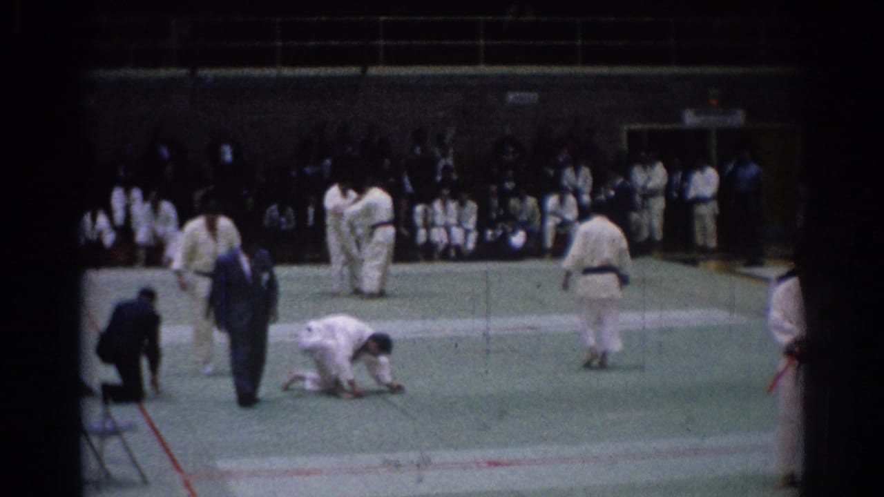1967 Los Angeles 8mm frame of karate athletes on a green auditorium mat with judges and spectators in the background.
