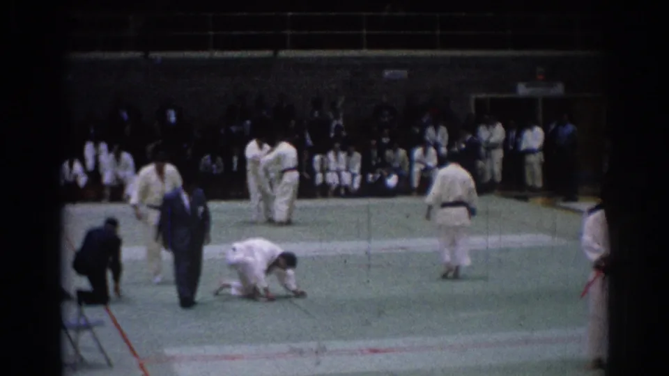 1967 Los Angeles 8mm frame of karate athletes on a green auditorium mat with judges and spectators in the background.