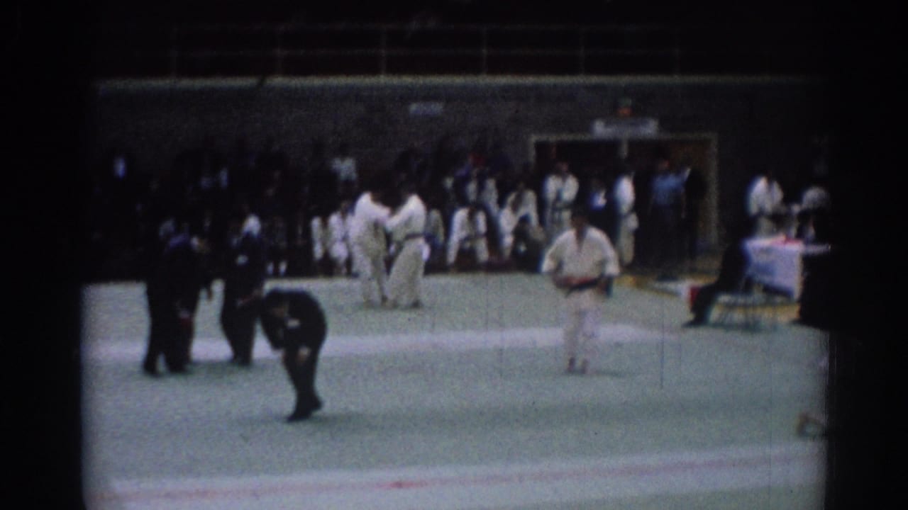 1967 Los Angeles karate competition frame showing two athletes on the mat, officials nearby, and spectators along the wall.