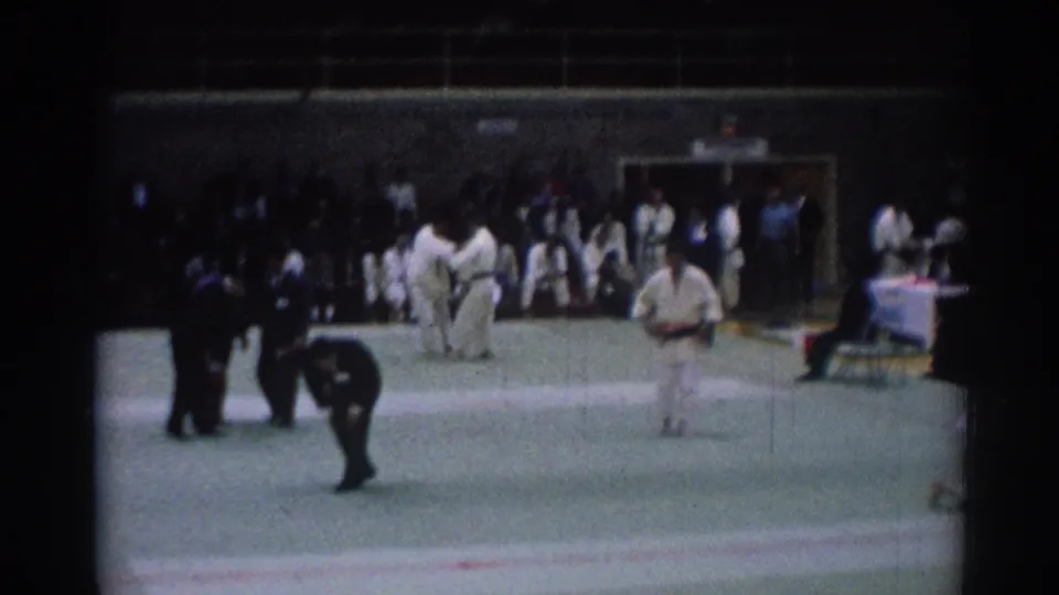 1967 Los Angeles karate competition frame showing two athletes on the mat, officials nearby, and spectators along the wall.