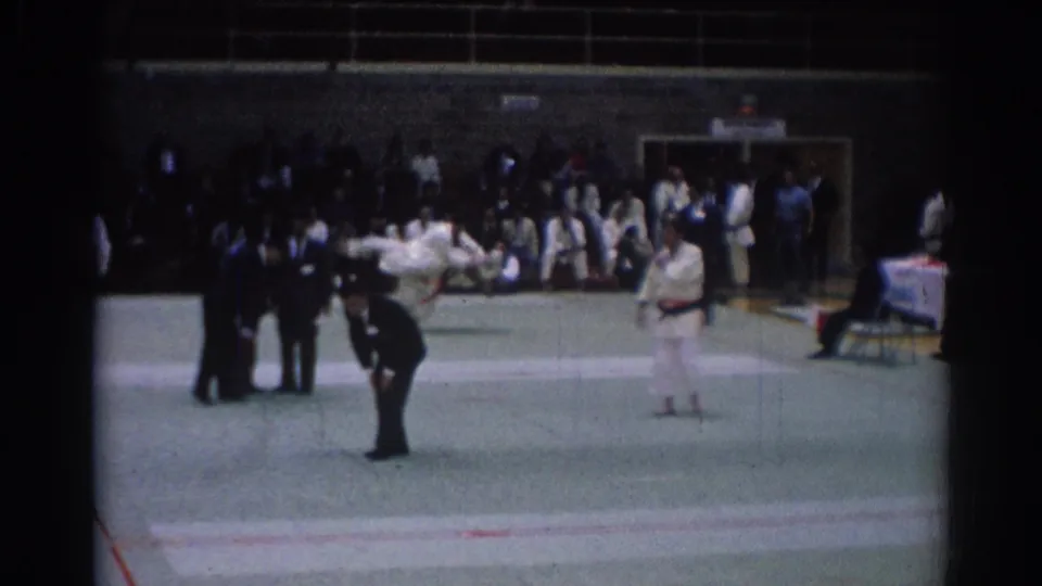 1967 Los Angeles home-movie frame of a karate match, with competitors in white gi uniforms, a suited referee, and spectators along the gym wall.