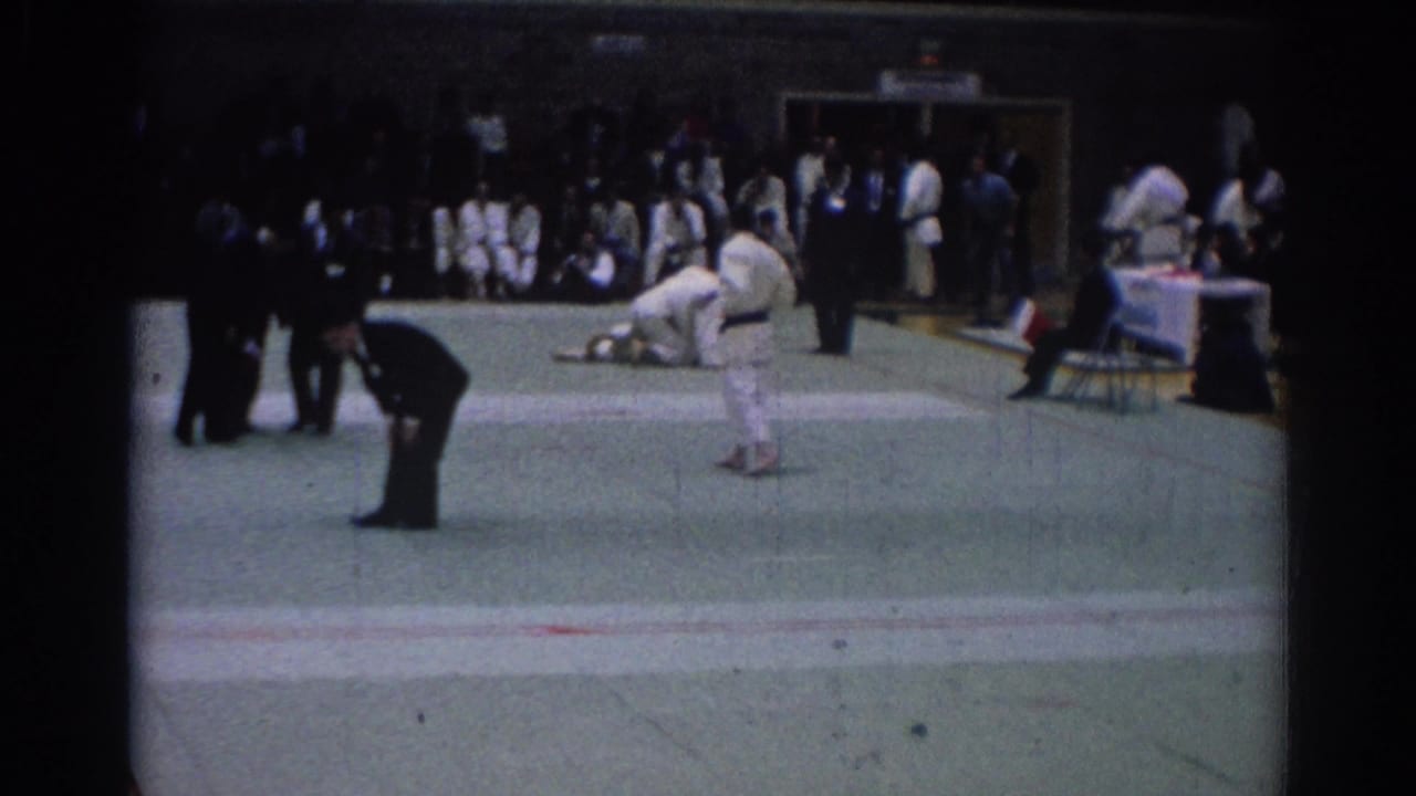 A 1967 Los Angeles karate bout appears mid-action, with one suited referee leaning in and a crowd watching from the edge of the gym floor.