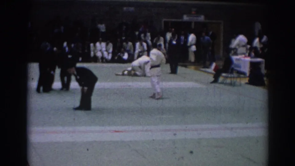 A 1967 Los Angeles karate bout appears mid-action, with one suited referee leaning in and a crowd watching from the edge of the gym floor.
