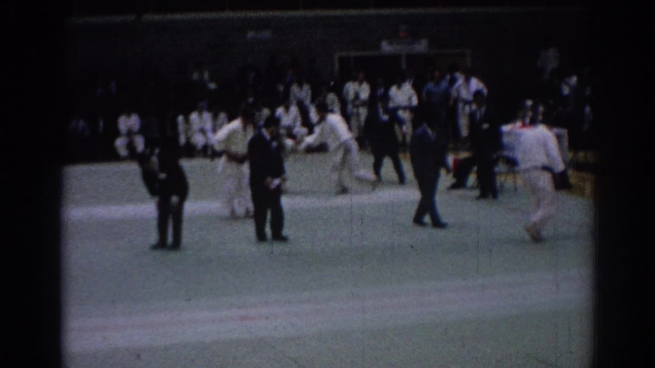 Indoor karate footage from 1967 Los Angeles shows competitors, suited officials, and spectators gathered around a green mat during a match exchange.
