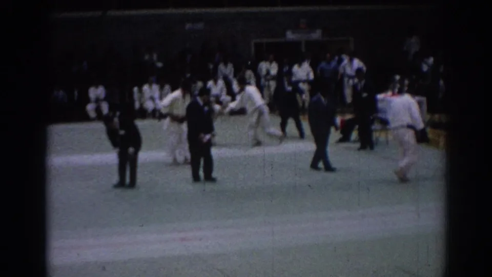 Indoor karate footage from 1967 Los Angeles shows competitors, suited officials, and spectators gathered around a green mat during a match exchange.