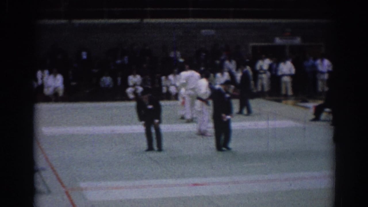A late frame from the 1967 Los Angeles karate event shows suited officials in front, competitors on the mat, and spectators lining the gym wall.