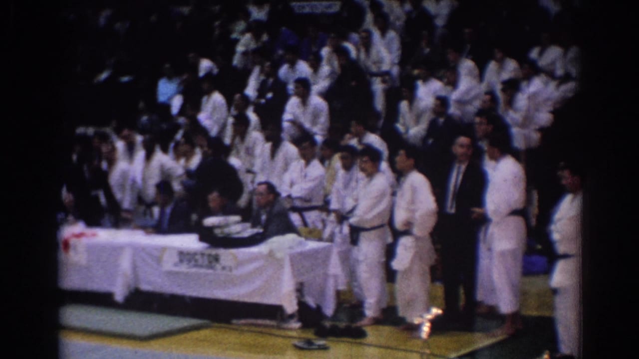 Karate competitors in white gi stand near officials seated at a table marked DOCTOR.