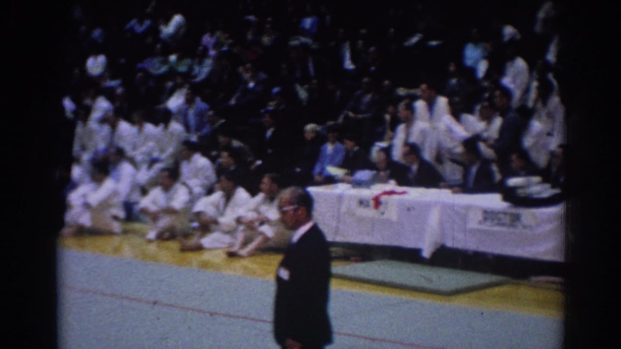 Indoor karate tournament view with a suited official in the foreground, seated white-gi participants, and a covered table in the background.