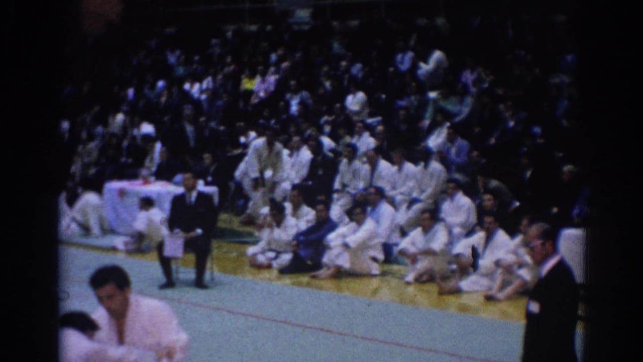 Wide 1967 Los Angeles karate tournament view with seated participants in white gi, spectators, gym mats, and a suited official at the edge.