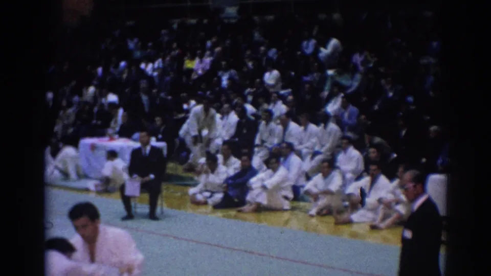 Wide 1967 Los Angeles karate tournament view with seated participants in white gi, spectators, gym mats, and a suited official at the edge.