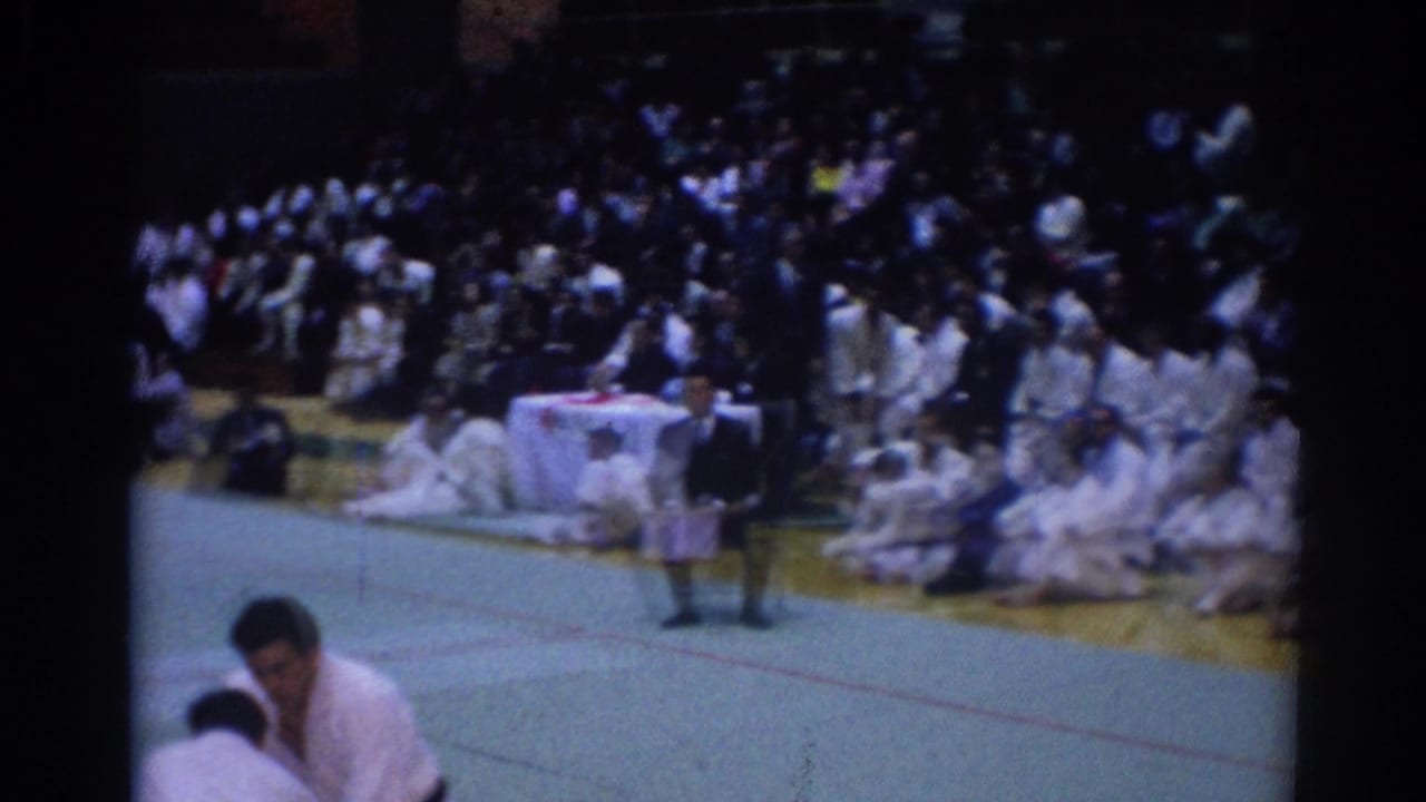 Judo competitors in white uniforms on a mat with spectators seated behind them.