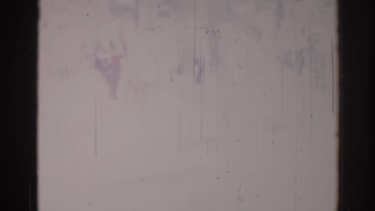 Pale damaged film frame with dark borders, vertical scratches, dust, and faint color staining.