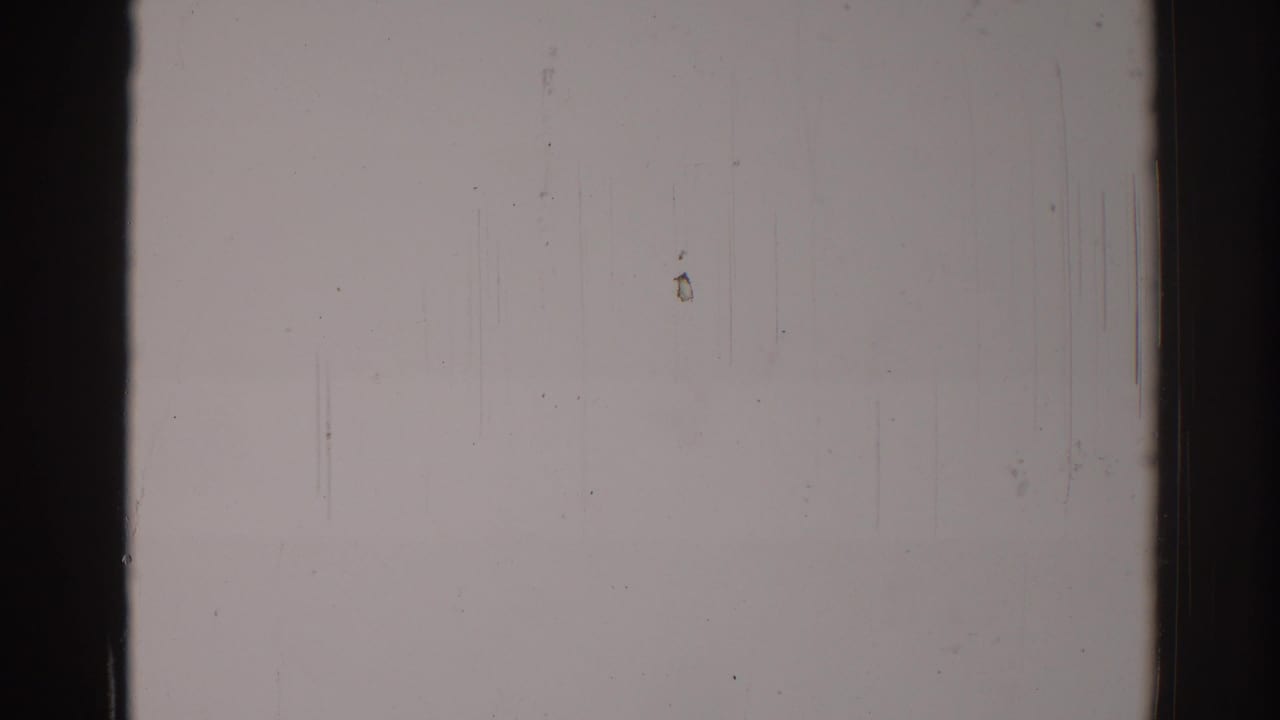 1967 Los Angeles 8mm frame of a blank pale wall with dark gate edges, scattered dust, fine scratches, and a small yellowish smudge.