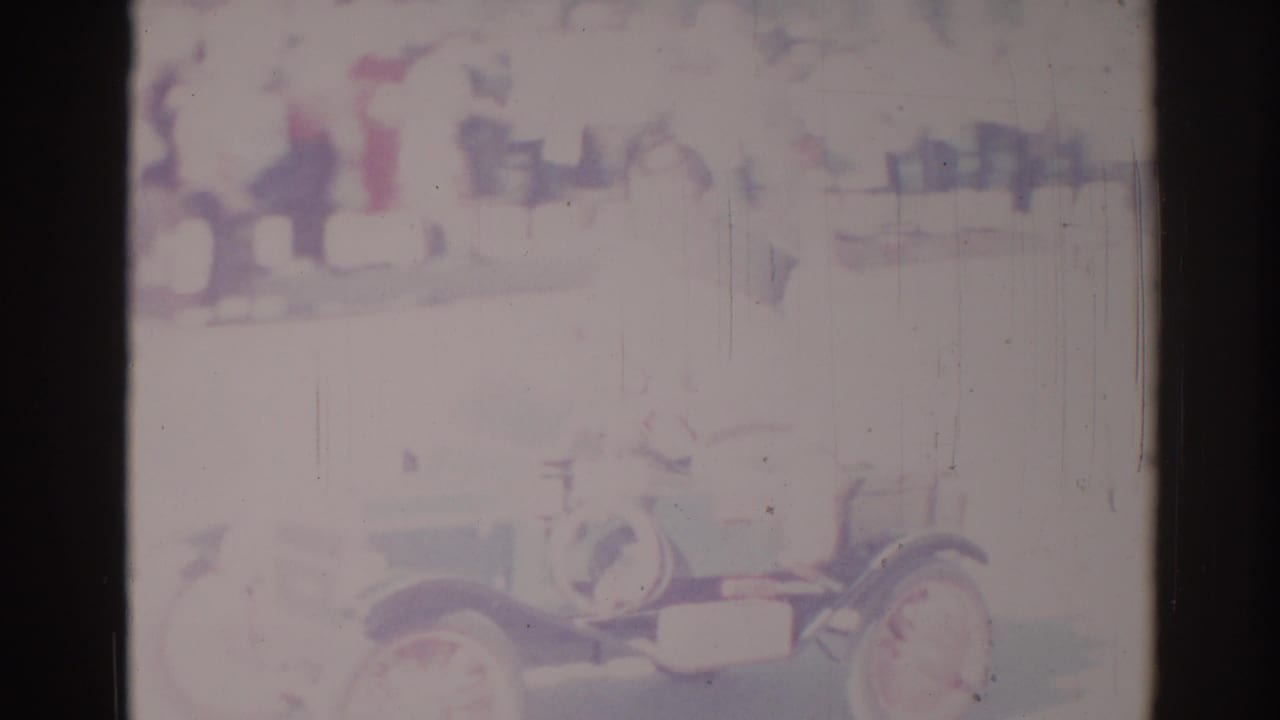 Faded parade frame with an antique-style open car and indistinct spectators along a street.