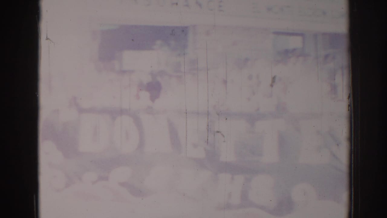 Blurred 1967 Los Angeles 8mm frame showing street-level storefront shapes, people, and a wide unreadable sign or banner.