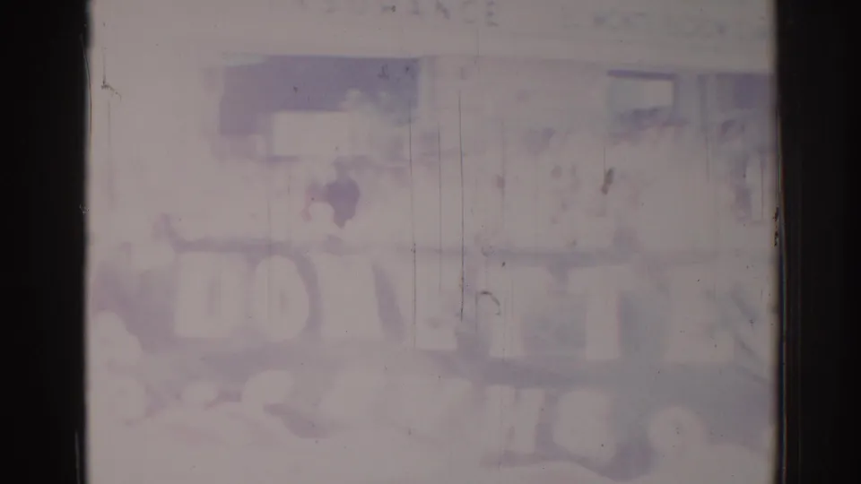 Blurred 1967 Los Angeles 8mm frame showing street-level storefront shapes, people, and a wide unreadable sign or banner.