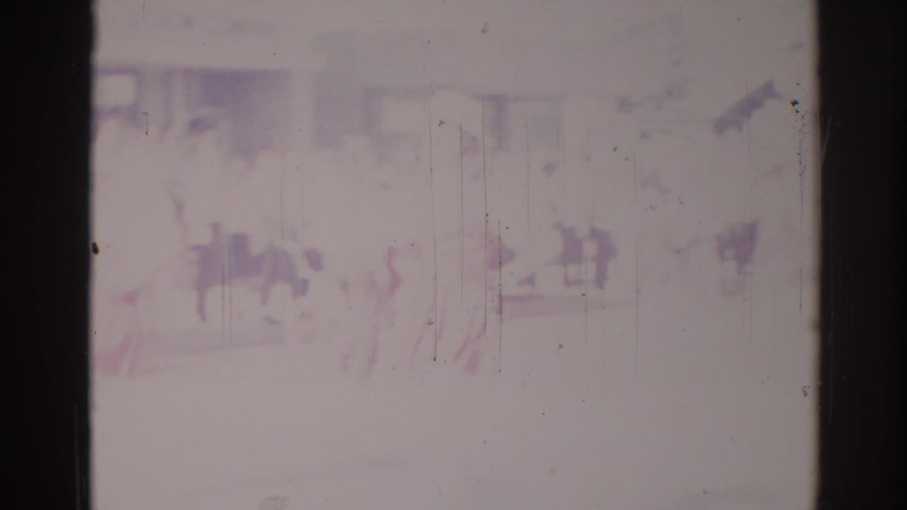 Faded 1967 Los Angeles parade footage shows indistinct people near a pale street-side building, framed by dark 8mm film edges.
