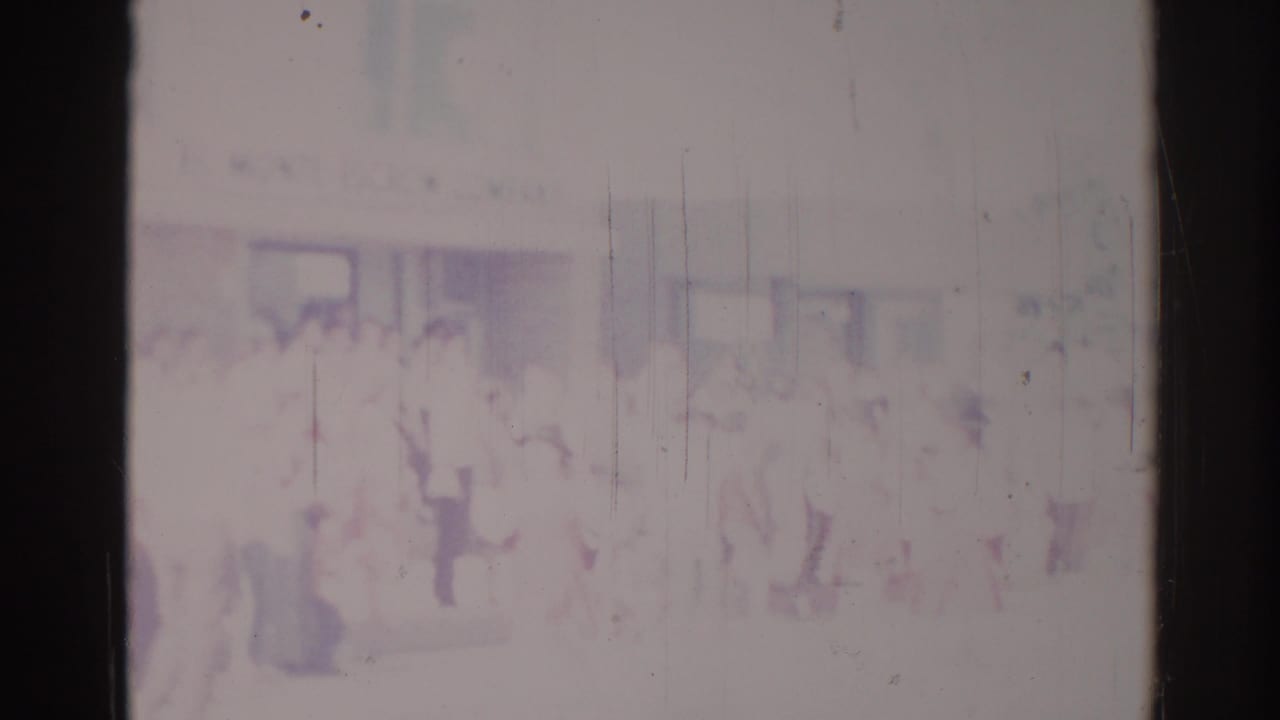 Blurred procession with faint standing figures and spectators against a pale building.