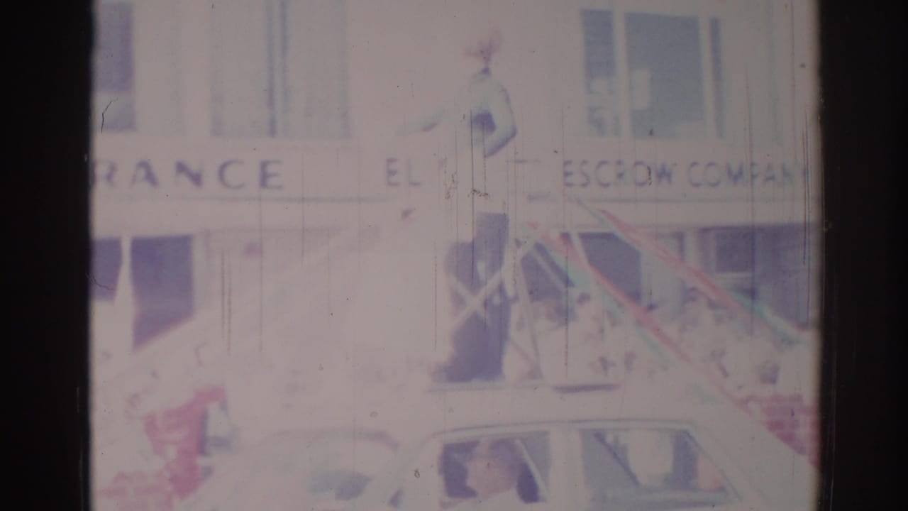 Faded street parade frame with a woman waving near storefront signage and a car below.
