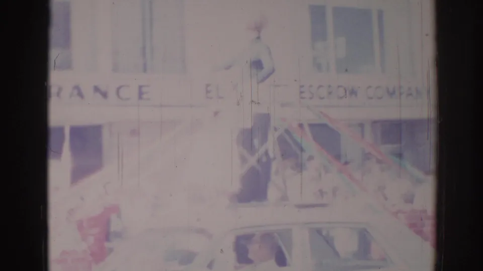 1967 Los Angeles-area 8mm frame showing a woman waving from an elevated parade position near escrow company signage and a car below.
