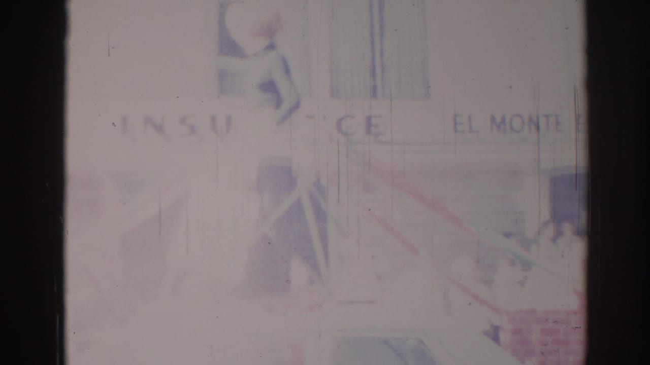 Faded 1967 Los Angeles-area street parade frame with insurance and El Monte signage, a car below, and indistinct spectators near the sidewalk.
