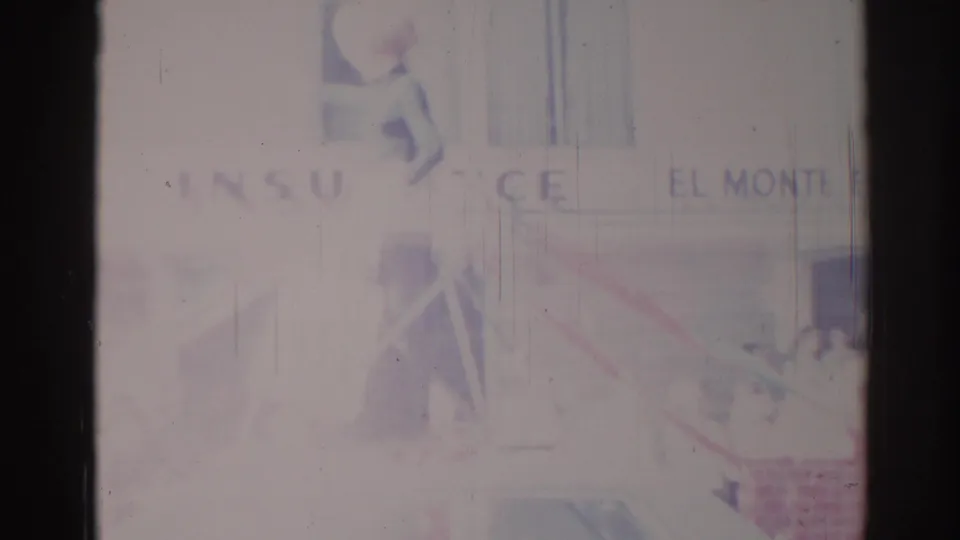 Faded 1967 Los Angeles-area street parade frame with insurance and El Monte signage, a car below, and indistinct spectators near the sidewalk.