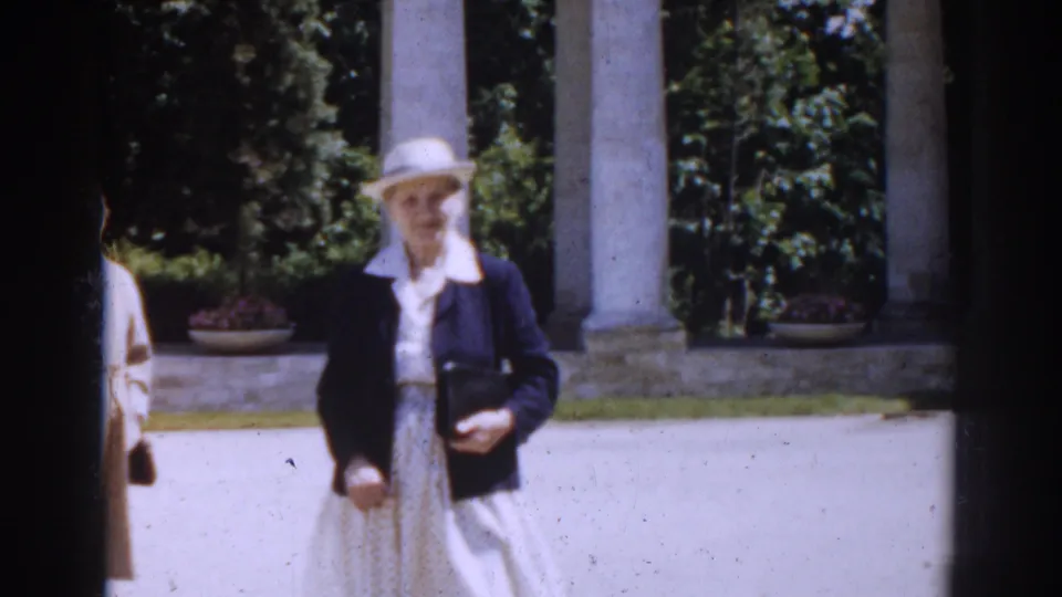 In 1968 Moscow, a woman in a white hat, dark jacket, and light patterned dress walks toward the camera before stone columns and greenery.