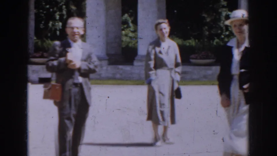 A suited man carrying a briefcase stands near a woman in a trench coat as another woman in a white hat enters the edge of the Moscow courtyard frame.