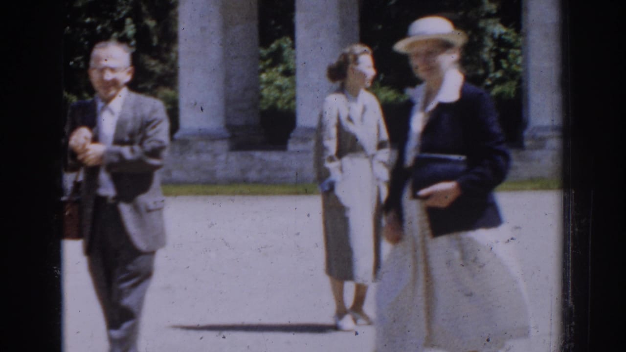 Three adults in a sunlit courtyard, with a man in a suit at left and two women near columns.