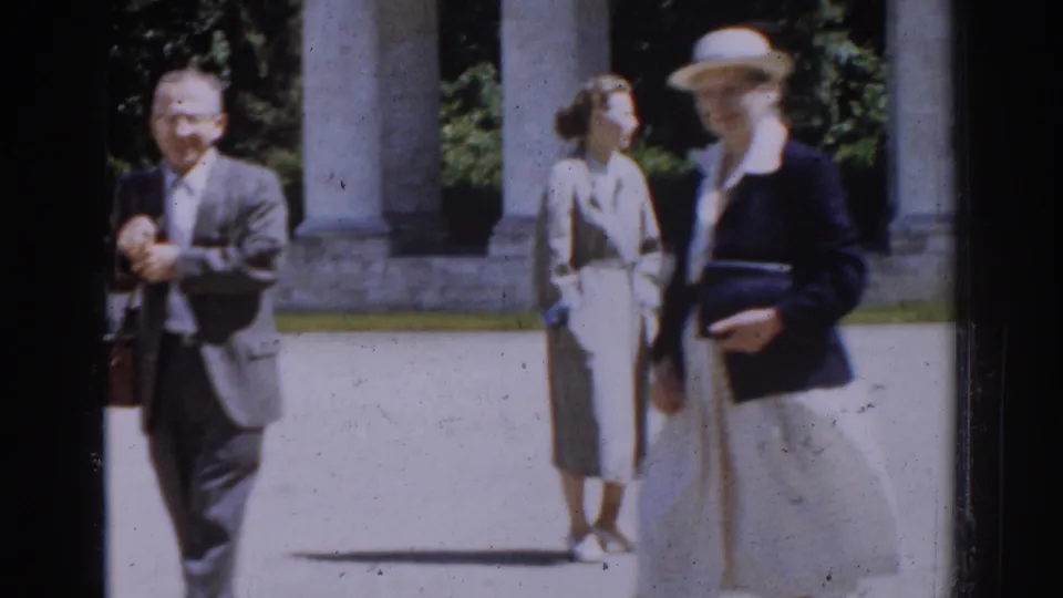 1968 Moscow home-movie frame showing adults in a bright courtyard near columns, including a suited man and two women in dresses or skirts.