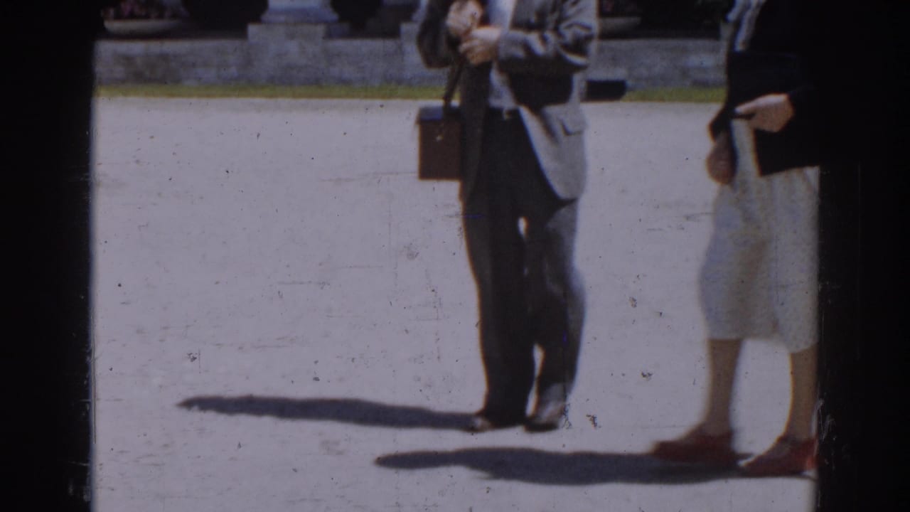 Lower bodies of adults walking across bright paving, with a briefcase and long shadows visible.