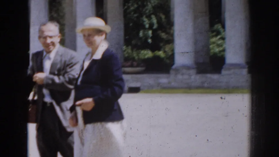 1968 Moscow frame of a suited man carrying a briefcase walking beside a woman in a brimmed hat, dark jacket, and light skirt.