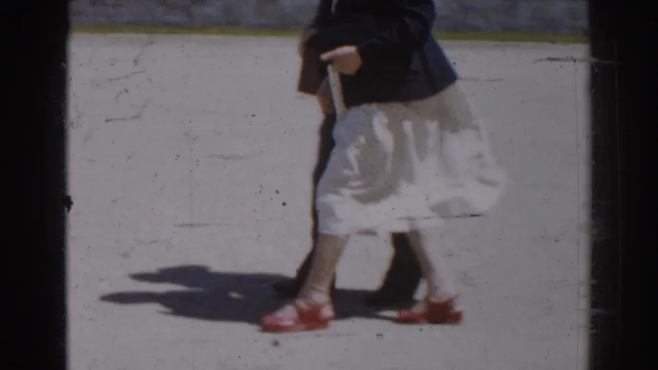 Cropped 1968 Moscow home-movie frame showing a woman’s light skirt, dark jacket, handbag, and red shoes while walking.
