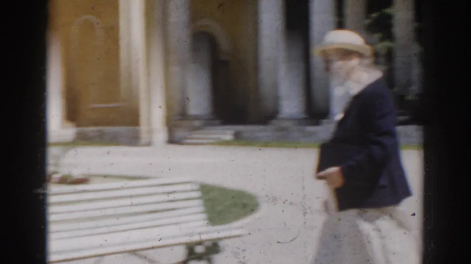 The woman continues across the Moscow garden setting, passing a white bench in front of columns and a yellow wall.