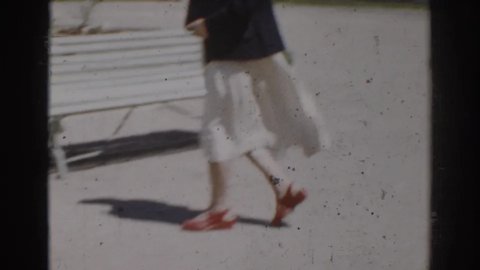 A wind-blown light skirt and red heels are visible as the woman walks across the frame near the white garden bench.