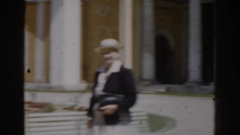 The woman appears to pause or slow near the white bench, with columns and a yellow building facade behind her.