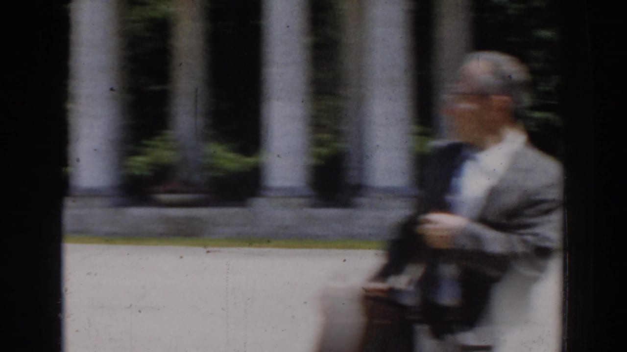 In 1968 Moscow home-movie footage, a suited man with glasses passes classical columns and greenery in soft focus.