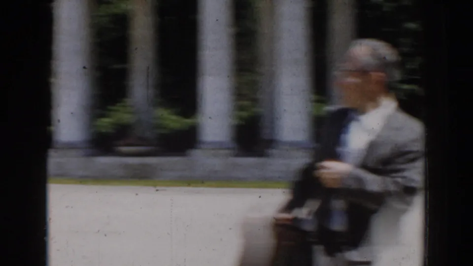 In 1968 Moscow home-movie footage, a suited man with glasses passes classical columns and greenery in soft focus.