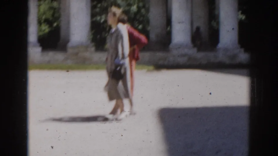 Two women in period dresses walk together through a Moscow park area with columns and foliage behind them.