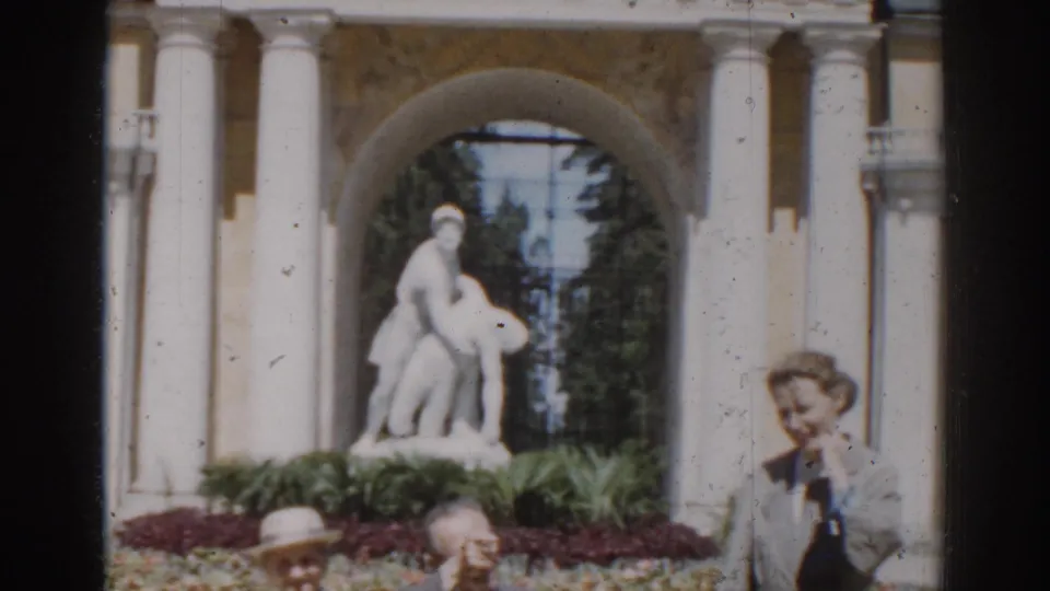 1968 Moscow home-movie frame showing an older couple seated near a standing woman, with a white statue and arched colonnade behind them.