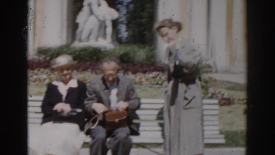 1968 Moscow home-movie frame of an older couple seated on a white bench, with a standing woman, flowerbeds, and a white statue behind them.