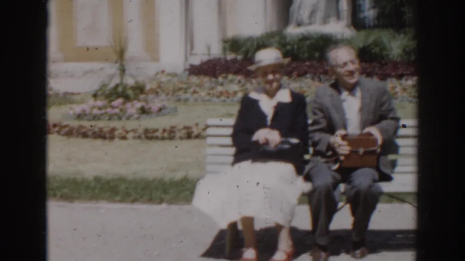 1968 Moscow frame showing an older couple seated side by side on a white bench in a manicured garden with pale columns and flowers behind them.